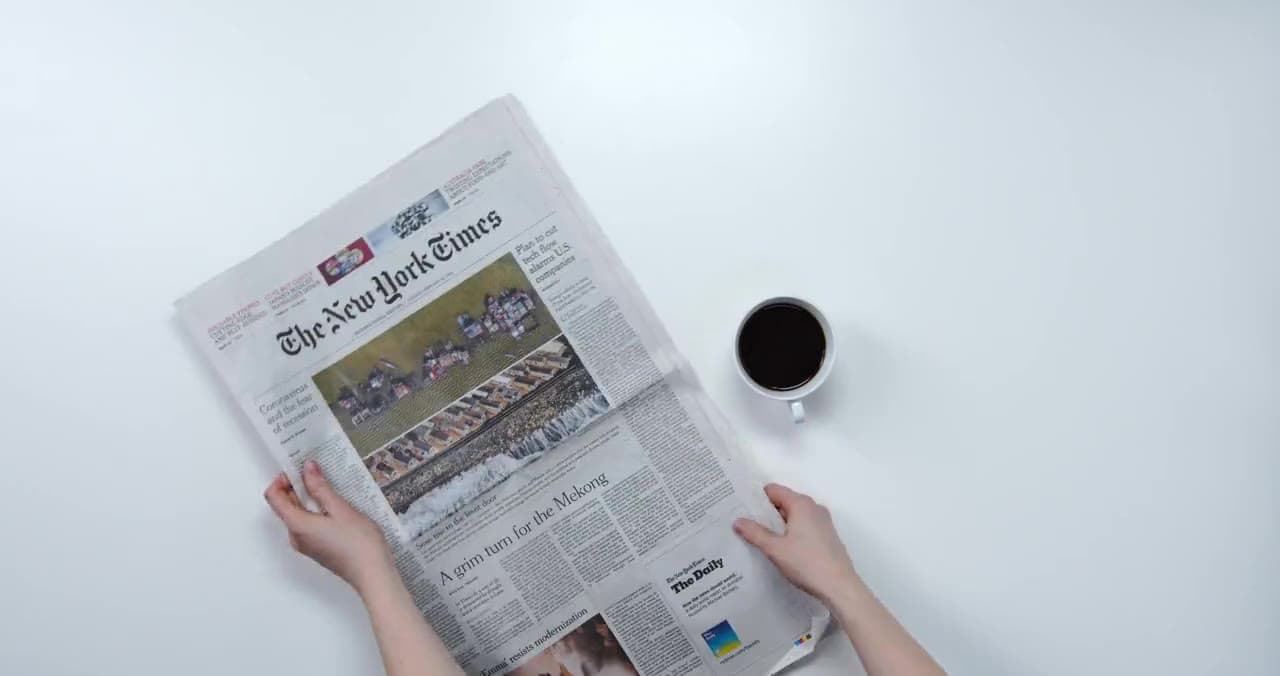 Hands holding an open New York Times newspaper beside a cup of black coffee on a white table.